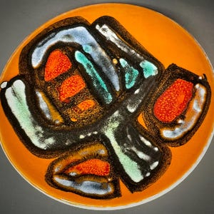 Puede incluir: Un plato redondo de cerámica naranja con un diseño abstracto único. El diseño presenta formas negras, blancas, azules y verdes con acentos naranjas y rojos texturizados. El plato tiene un acabado brillante y un borde blanco.