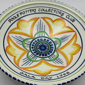 May include: A white Poole Pottery Collectors Club plate with a floral design in the center. The flower has yellow, orange, and blue accents. The plate has a blue and green border, with the words "Gala Day 1996" around the edge.