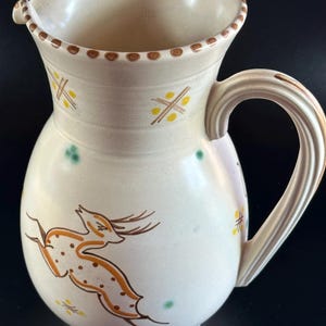 May include: A white ceramic pitcher with a brown handle and a stylized deer design in brown, yellow, and green. The pitcher has a brown rim with a row of brown dots.