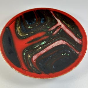 Puede incluir: Un plato redondo de cerámica con un borde rojo vibrante y un diseño interior llamativo. El interior presenta un patrón arremolinado de negro, rojo y toques de verde y dorado. El diseño del plato es abstracto y atractivo.