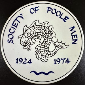 May include: A white ceramic plate with a black and blue design. The plate features a stylized fish illustration in black, surrounded by the text "SOCIETY OF POOLE MEN" in blue. The years "1924" and "1974" are also present.