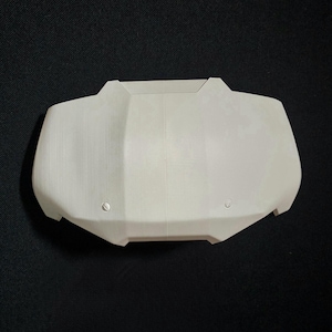 May include: A light-colored, geometric object with a symmetrical design. The object has a smooth surface with two small, circular indentations near the bottom. The overall shape is rectangular with angled edges and a central ridge.