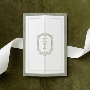 May include: A rectangular gatefold invitation card featuring a white center panel with a sage green ornate frame and the letters E & J in the middle. The card has sage green outer panels and is accented by a long, flowing white satin ribbon.