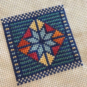 Può includere: Un motivo a punto croce colorato con un design blu, verde acqua, rosso, arancione e giallo. Il motivo è ricamato su tela Aida bianca.