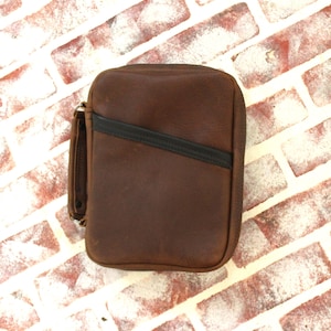 May include: A rectangular brown leather case with a smooth matte finish and a dark brown diagonal accent stripe across the front. The case features a sturdy side handle and a zipper closure that runs along the top and sides of the structure.