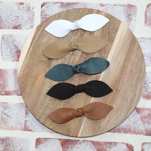 Puede incluir: Cinco pinzas para el pelo de cuero en forma de lazo de diferentes colores sobre una superficie de madera. Los colores son blanco, marrón, azul verdoso, negro y marrón.