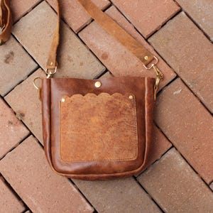 Puede incluir: Un bolso de cuero marrón cruzado con cierre de solapa y un bolsillo festoneado. El bolso tiene una hebilla y herrajes dorados.