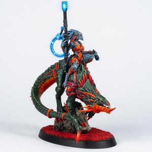 May include: A detailed miniature figure of a warrior riding a reptilian creature. The warrior is clad in orange and blue armor, holding a glowing staff. The creature is green and red, with sharp details. The base is oval and covered in red flocking.