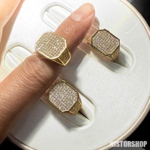 Peut inclure: Bague dorée avec une face rectangulaire recouverte de petites pierres scintillantes. La bague est présentée sur un doigt et dans un présentoir à bijoux. La bague a un anneau détaillé et texturé.