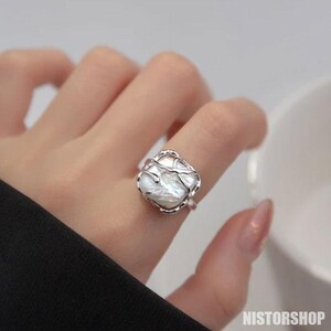 Peut inclure: Bague en argent ornée d'une grosse perle blanche de forme irrégulière. La perle est sertie dans un délicat entourage ajouré en argent. La bague est portée au doigt, avec une manche noire en arrière-plan.