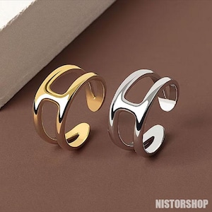 Peut inclure: Deux bagues réglables, une dorée et une argentée, sont présentées côte à côte. Chaque bague présente un design à double bande avec une forme ouverte et incurvée. Les bagues sont exposées sur une surface marron. Le texte "NISTORSHOP" est en bas à droite.