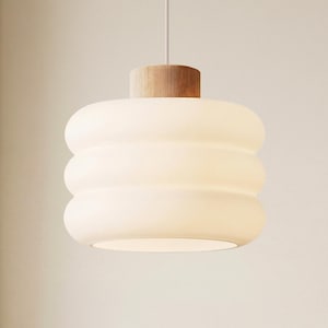 Op de afbeelding: Een moderne hanglamp met een cilindrisch, gestapeld ontwerp. De lamp heeft een houten bovenkant en een matglazen kap. Daaronder staat een houten tafel met een kleine, ronde, bruine kom in een grotere, gebroken witte kom.
