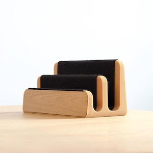 May include: A tiered desk organiser featuring a light-coloured wood base with a ribbed side texture and two black felt-lined slots for holding items. The structure has a stepped design with a taller back section and a shorter front section.
