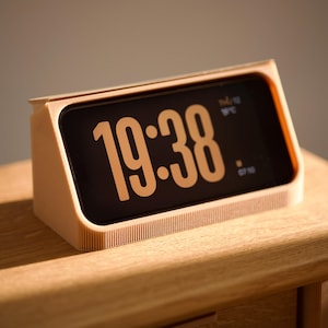 Può includere: Una sveglia digitale con cornice beige mostra l'ora 19:38 in cifre arancioni su uno schermo nero. L'orologio è appoggiato su una superficie di legno, con l'ora e la temperatura visibili.