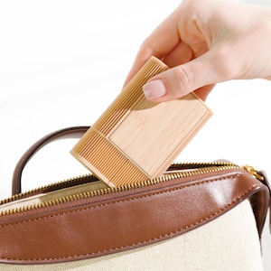 Puede incluir: Una caja de madera de color claro con un diseño acanalado se coloca en un bolso beige y marrón. La caja tiene forma rectangular con una superficie lisa y plana y una sección texturizada y acanalada. El bolso tiene cierre de cremallera.