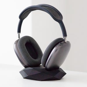 May include: Dark gray over-ear headphones resting on a black geometric stand. The headphones have a padded headband and earcups. The stand has a faceted design. The background is white.