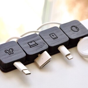 Puede incluir: Un organizador de cables de madera negra con cuatro secciones cuadradas, cada una con un icono diferente: auriculares, un port&aacute;til, una tableta y un reloj inteligente. Cables y conectores de carga blancos enchufados.