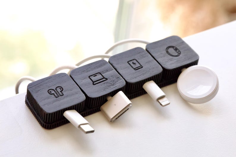 Puede incluir: Un organizador de cables de madera negra con cuatro secciones cuadradas, cada una con un icono diferente: auriculares, un port&aacute;til, una tableta y un reloj inteligente. Cables y conectores de carga blancos enchufados.
