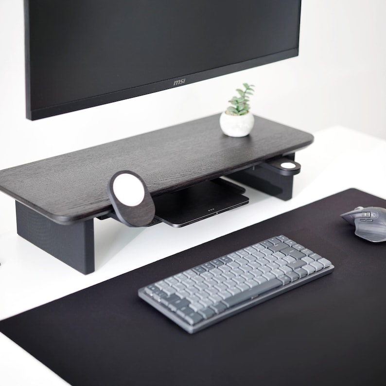 May include: A dark wood-tone desk riser with a small plant in a white pot. A black keyboard, mouse, and mousepad are on the desk. A black monitor is mounted above the desk riser.