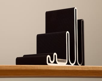 May include: A modern black bookend with a unique, stepped design. The bookend features a series of stacked, rectangular forms with a textured surface and white accents. It sits on a wooden shelf against a neutral background.