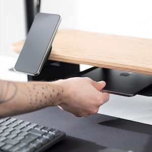 May include: A wooden desk organizer with a light wood finish, featuring a built-in shelf and a black smartphone holder. A black tablet is being placed on the shelf. A keyboard is visible in the foreground.