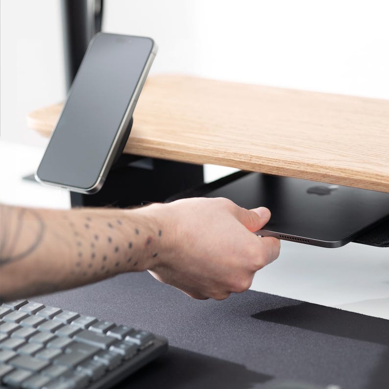 May include: A wooden desk organizer with a light wood finish, featuring a built-in shelf and a black smartphone holder. A black tablet is being placed on the shelf. A keyboard is visible in the foreground.