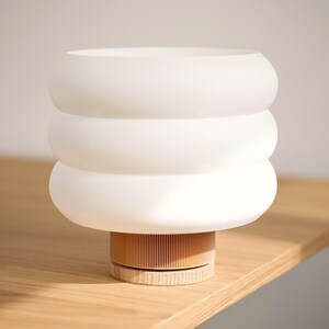 Puede incluir: Una l&aacute;mpara de mesa con una pantalla de vidrio esmerilado blanco de tres niveles redondeados. La base consta de un componente cil&iacute;ndrico de color marr&oacute;n claro con estr&iacute;as verticales sobre un soporte de madera.