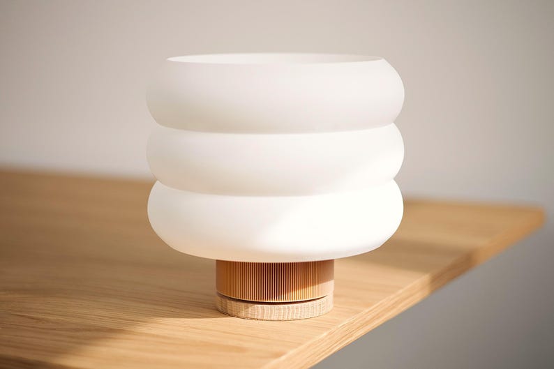 Puede incluir: Una l&aacute;mpara de mesa con una pantalla de vidrio esmerilado blanco de tres niveles redondeados. La base consta de un componente cil&iacute;ndrico de color marr&oacute;n claro con estr&iacute;as verticales sobre un soporte de madera.