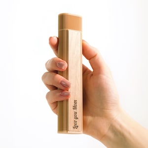 May include: A light brown wooden rectangular box with a ribbed top and a smooth, flat side. The text "Love you Mom" is engraved on the side. The box is held in a person's hand against a white background.