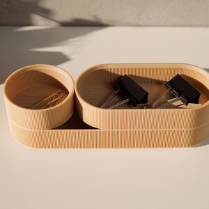 May include: A set of two wooden desktop organizers with a natural finish. The larger organizer is oval shaped and has a compartment for holding paper clips. The smaller organizer is round and has a compartment for holding paper clips.