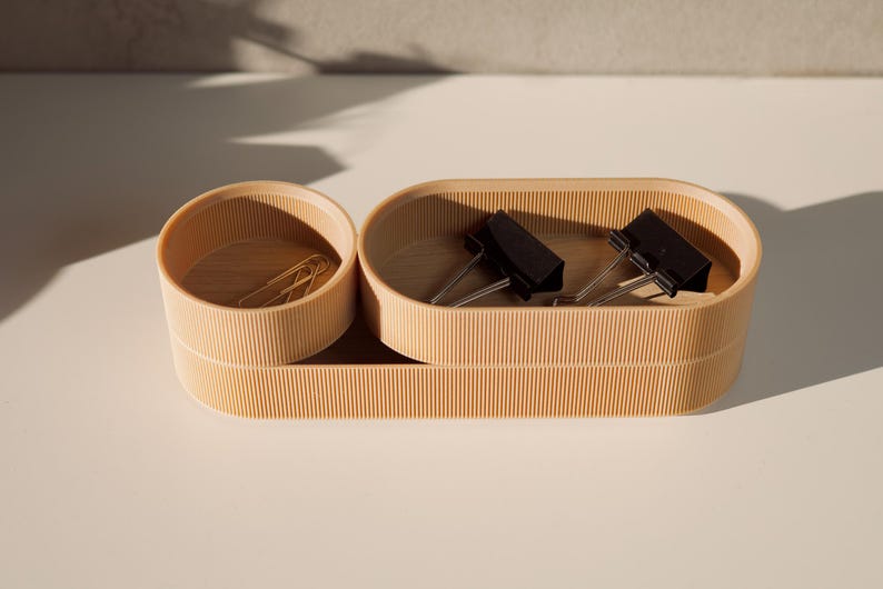 May include: A set of two wooden desktop organizers with a natural finish. The larger organizer is oval shaped and has a compartment for holding paper clips. The smaller organizer is round and has a compartment for holding paper clips.