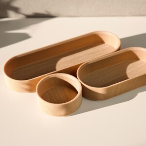 May include: Three wooden oval-shaped trays with a ribbed texture. The trays are nested inside each other, with the largest tray at the bottom and the smallest tray at the top.