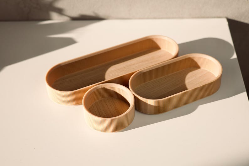 May include: Three wooden oval-shaped trays with a ribbed texture. The trays are nested inside each other, with the largest tray at the bottom and the smallest tray at the top.