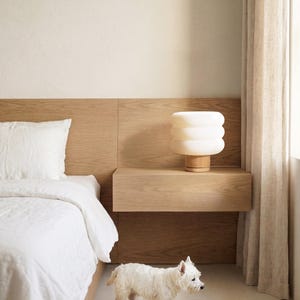 Puede incluir: Una escena de dormitorio con una cama blanca con almohada y edred&oacute;n a juego. Una l&aacute;mpara moderna blanca apilada se encuentra sobre una mesita de noche de madera. Un perro peque&ntilde;o blanco camina por el suelo. La habitaci&oacute;n tiene paredes claras y una paleta de colores neutros.