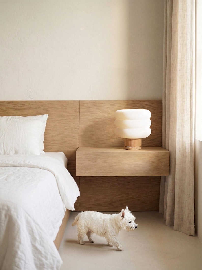 Puede incluir: Una escena de dormitorio con una cama blanca con almohada y edred&oacute;n a juego. Una l&aacute;mpara moderna blanca apilada se encuentra sobre una mesita de noche de madera. Un perro peque&ntilde;o blanco camina por el suelo. La habitaci&oacute;n tiene paredes claras y una paleta de colores neutros.