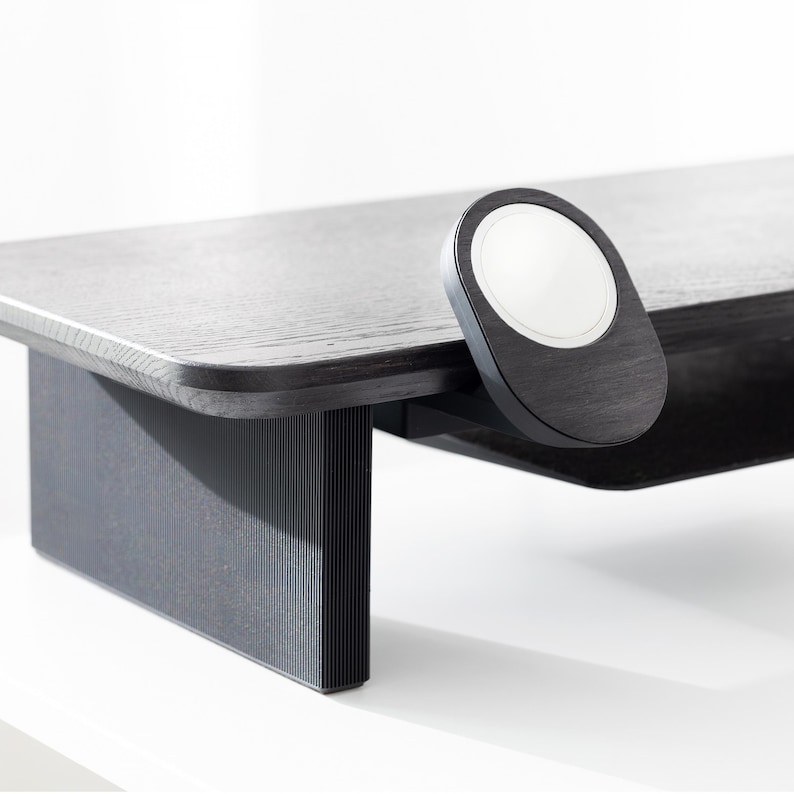 May include: A dark wood desk with a rounded edge and a black, oval-shaped wireless charger with a white center. The desk has a matching dark wood leg with vertical lines.