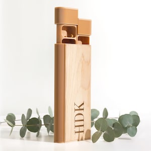 May include: A vertical wooden pill organizer with a light tan finish and ribbed texture on the top and side panels. The front features the engraved letters HDK. The top section has two open compartments, one containing small pills, with matching tan lids.