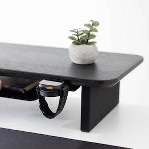 May include: A black wooden desk with a built-in shelf and a black watch with a mesh band hanging from the edge. A small potted plant sits on the desk's surface. The desk has a modern design and a minimalist aesthetic.