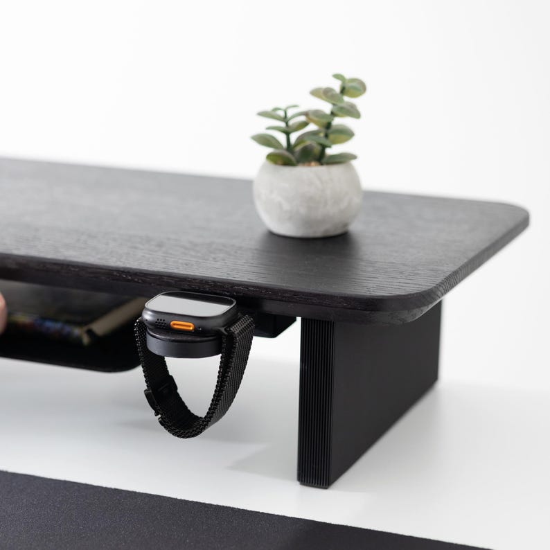 May include: A black wooden desk with a built-in shelf and a black watch with a mesh band hanging from the edge. A small potted plant sits on the desk's surface. The desk has a modern design and a minimalist aesthetic.