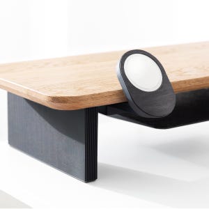 May include: A wooden monitor stand with a black base and a black, oval-shaped wireless charger attached to the side. The stand has a light brown, rectangular top and a black, grooved base. The wireless charger has a white charging surface.