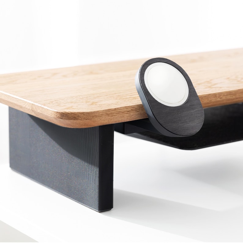 May include: A wooden monitor stand with a black base and a black, oval-shaped wireless charger attached to the side. The stand has a light brown, rectangular top and a black, grooved base. The wireless charger has a white charging surface.