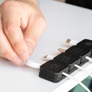 Puede incluir: Un organizador de cables negro con cuatro ranuras, cada una con un cable blanco con un conector diferente. El organizador est&aacute; sobre una superficie blanca y una mano sostiene uno de los cables.