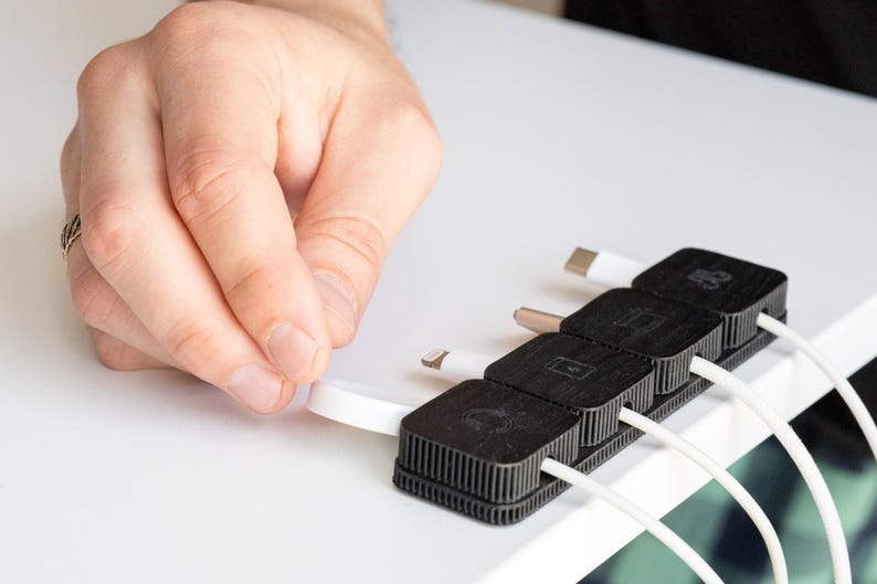 Puede incluir: Un organizador de cables negro con cuatro ranuras, cada una con un cable blanco con un conector diferente. El organizador est&aacute; sobre una superficie blanca y una mano sostiene uno de los cables.