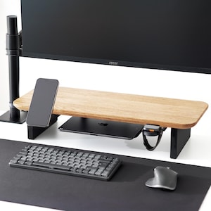 May include: A wooden monitor stand with a light brown surface and black legs. A smartphone, a tablet, and a smartwatch are on the stand. A keyboard and mouse are on a black desk mat in the foreground. A black monitor is in the background.