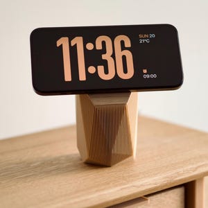 May include: A black smartphone with a wooden stand displaying the time 11:36, the date SUN 20, and the temperature 21°C. The stand is made of light brown wood with a geometric design.