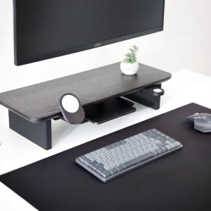 May include: A dark wood-tone monitor stand with a built-in shelf and a small white plant in a pot. A black keyboard, mouse, and mousepad are on the desk. The monitor is black.