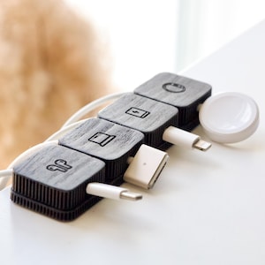 Puede incluir: Un organizador de cables de madera gris oscuro con cuatro secciones cuadradas, cada una con un icono diferente. Cables de carga blancos y un cargador de Apple Watch blanco están conectados. El organizador está sobre una superficie blanca.