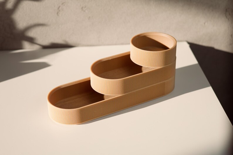 May include: Three stacked oval-shaped containers made of a light brown material with a ribbed texture. The containers are nested inside each other, creating a tiered effect.
