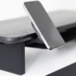 May include: A black wooden monitor stand with a built-in wireless charger. A silver and black smartphone is charging on the stand. The stand has a dark wood grain finish and a black base.