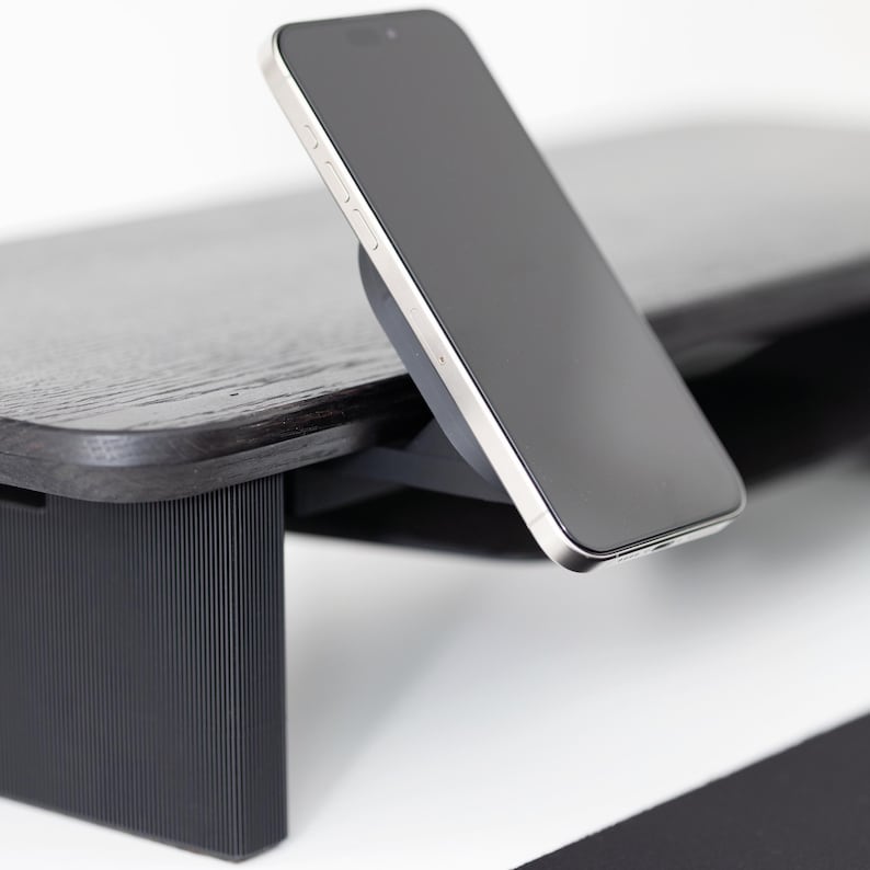 May include: A black wooden monitor stand with a built-in wireless charger. A silver and black smartphone is charging on the stand. The stand has a dark wood grain finish and a black base.